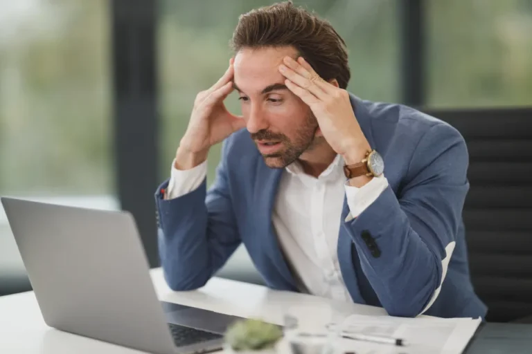 A businessman at his desk holding his head in frustration in front of a laptop