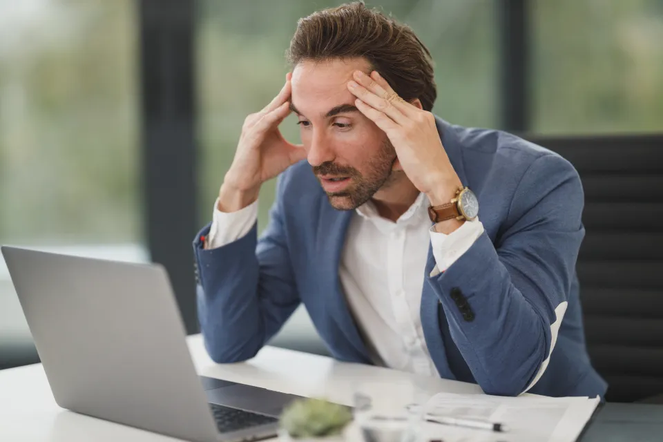 A businessman at his desk holding his head in frustration in front of a laptop